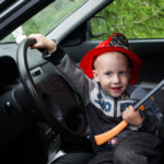 baby boy firefighter fireman suit, smiling, car, wheel, driver, helmet