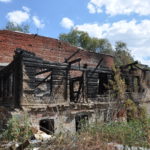 Abandoned burnt house of old architecture with ruined wooden and brick walls and charry logs, all among green trees, bushes and garbage. Big clouds in the sky.